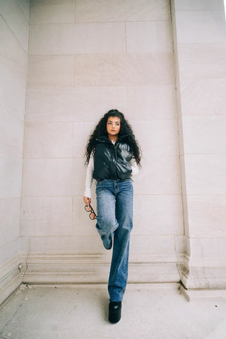 girl leaning against a building wearing a zipped faux leather puffer vest and wide leg denim