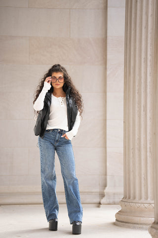 Chica con cabello rizado con gafas de sol, chaleco hinchado de cuero sintético y jeans anchos azules