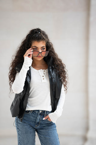 Una chica con el pelo largo y rizado que llevaba un chaleco bomber de piel sintética, una camiseta blanca de manga larga y gafas de sol.